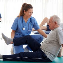 A young female physical therapist wearing blue scrubs and a stethoscope smiles while assisting an elderly man with a leg exercise on a green mat in a bright rehabilitation center, with other senior.